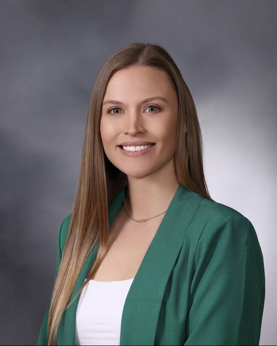 Haley Marsicek, with long, straight hair, smiles at the camera. She is wearing a white top and a green blazer. The background is a smooth gradient of gray tones, creating an elegantly understated backdrop for AINS's latest campaign photo.