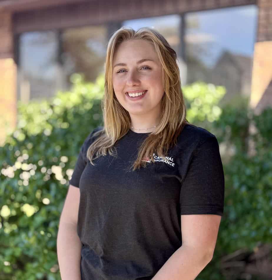 Amber Crocker, a young woman with blonde hair, smiles outdoors in front of green bushes and a building. She is wearing a black t-shirt and standing in bright natural light.